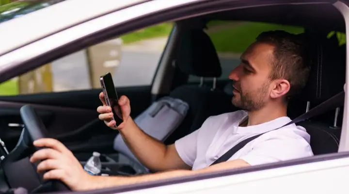 En medio del tráfico, un conductor mira un teléfono inteligente mientras está sentado en un automóvil estacionado, con una mano apoyada en el volante y un bolso en el asiento del pasajero.