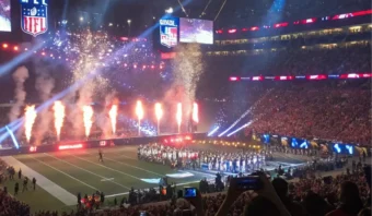Estadio de fútbol con fuegos artificiales y público durante un evento. Los participantes se reúnen en el campo, rodeados de luces y espectáculos, mientras los espectadores capturan el momento con sus teléfonos.