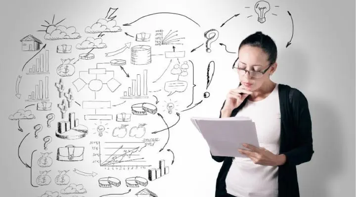 Una mujer examina un documento, de pie frente a una pared adornada con diversos dibujos y símbolos vinculados a los negocios y al análisis de datos, que reflejan ideas de 6 entidades líderes en emprendimiento.