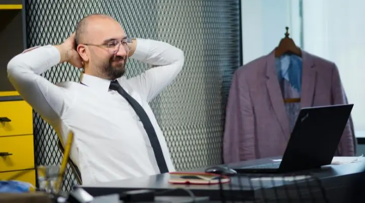 Un hombre con camisa blanca y corbata está sentado en un escritorio, con las manos detrás de la cabeza, sonriendo mientras analiza ideas sobre sostenibilidad humana. Una chaqueta rosa cuelga al fondo.