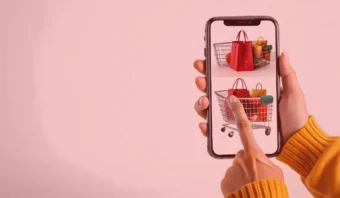 Una persona que lleva un jersey amarillo utiliza un teléfono inteligente para realizar compras en línea. La pantalla muestra imágenes de carritos de la compra llenos de varias bolsas de la compra.