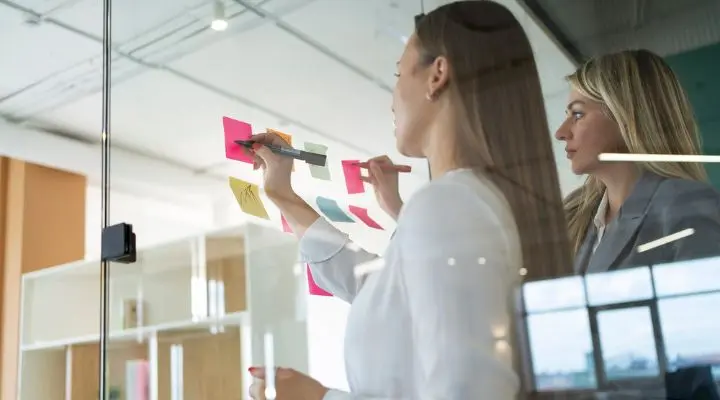En tiempos de cambio, dos personas en una oficina escriben en notas adhesivas de colores colocadas sobre una pared de cristal. Una usa un bolígrafo mientras la otra observa, capturando ideas e inspiración.