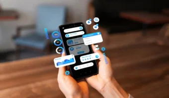 Persona que sostiene un teléfono inteligente que muestra varias pantallas e íconos digitales flotantes, incluidos gráficos, barras de búsqueda, notificaciones y configuraciones, sobre un fondo interior borroso.