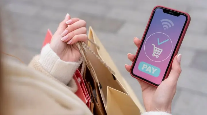 Una persona que sostiene bolsas de compras usa un teléfono inteligente con una interfaz de aplicación de pago móvil que muestra un ícono de carrito de compras y un botón "PAGAR".