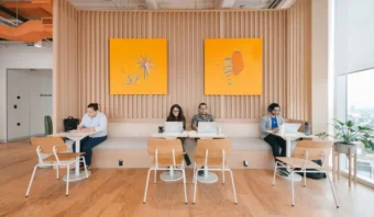 Cuatro personas trabajan con portátiles en mesas en una habitación moderna con detalles de madera. Dos cuadros amarillos brillantes cuelgan de la pared.