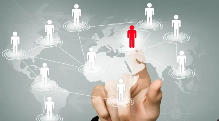 Una mano señala un mapa digital con figuras blancas interconectadas. Entre ellas, una figura es roja, lo que simboliza los stakeholders y la concentración o la selección en una red de actores a lo largo de los continentes.