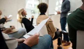 Personas sentadas, con papeles y bolígrafos en la mano, escuchando atentamente una presentación en una sala luminosa.