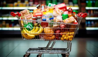 Un carrito de compras lleno de diversos comestibles, incluidos plátanos, bocadillos envasados, productos enlatados y productos frescos, en el pasillo de un supermercado.