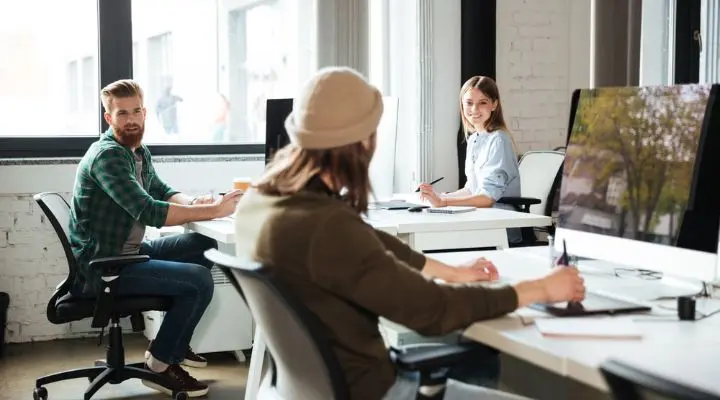 Tres personas sentadas en sus escritorios en una oficina moderna con grandes ventanales y computadoras. Un hombre con barba observa a una mujer sonriente, ejemplificando sus métodos de trabajo colaborativos, mientras otra persona se concentra atentamente en una tableta.