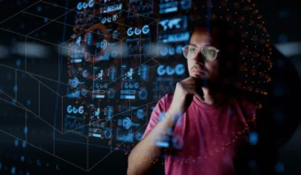 Un hombre con gafas y camisa roja mira pensativamente las pantallas digitales iluminadas que muestran datos y gráficos.