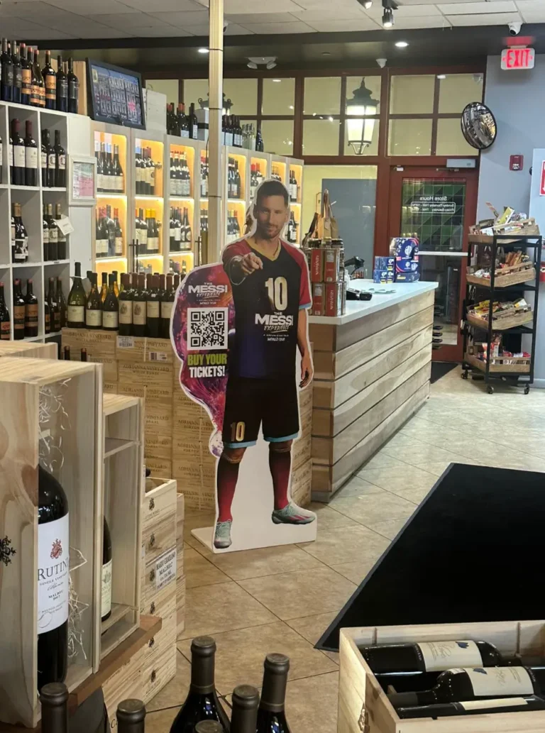 Un recorte de cartón de un jugador de fútbol añade un toque de estilo deportivo al ambiente de la tienda de vinos, junto al mostrador de madera en medio de estantes de botellas y, como un elemento OOH innovador, presenta un código QR para acceder fácilmente a las entradas.