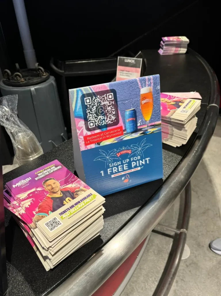 Pilas de folletos promocionales y un stand que anuncia "1 pinta gratis" con un código QR están colocados estratégicamente en un mostrador curvo, similar a un expositor OOH, junto a un portavasos de plástico en un entorno interior.