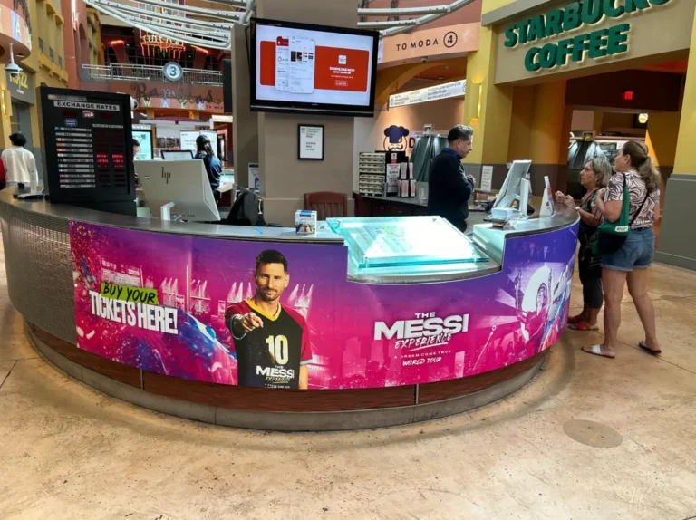 El mostrador de venta de entradas del centro comercial, decorado con publicidad exterior con temática de Messi, está convenientemente ubicado cerca de un café Starbucks. La gente interactúa con el empleado mientras las pantallas digitales y los paneles promocionales captan su atención.