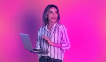 Una persona con una camisa a rayas sostiene una computadora portátil y sonríe contra un fondo degradado de color rosa y morado.