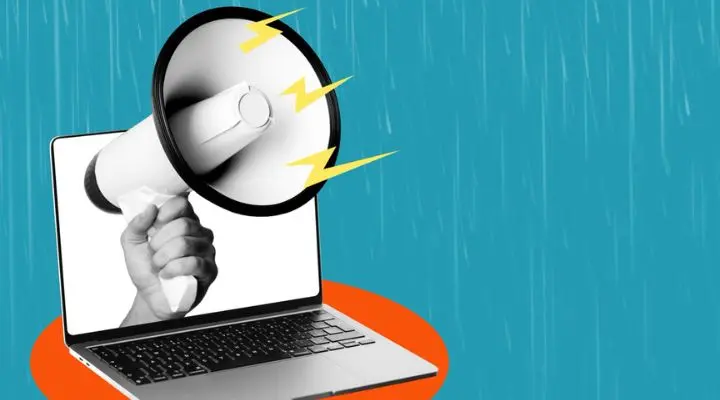 Una mano que sostiene un megáfono emerge de la pantalla de una computadora portátil sobre un fondo azul, simbolizando los anuncios en línea y resaltando el impacto de la planificación de medios en las estrategias de comunicación digital.