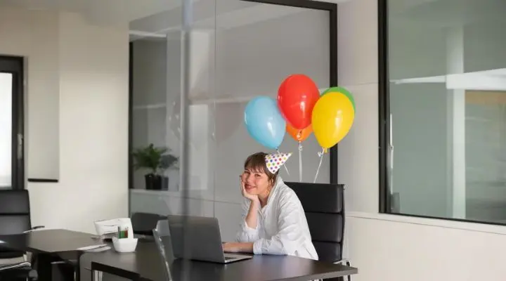 Una persona en una oficina con un sombrero de fiesta está sentada en un escritorio con una computadora portátil, rodeada de globos vibrantes atados a la silla: una escena perfecta para birthday marketing .