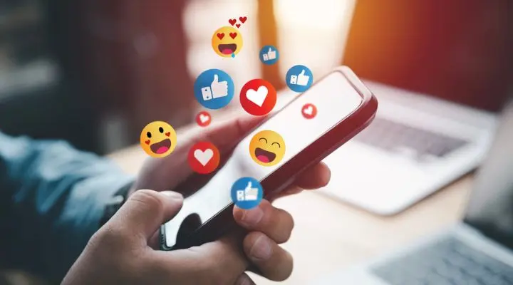 Una persona sostiene un teléfono inteligente mientras los íconos de reacción de las redes sociales, como corazones y me gusta, flotan sobre la pantalla, ilustrando el impacto de una meticulosa planificación de medios.