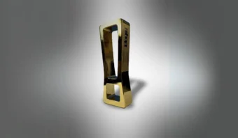Escultura rectangular de color dorado con un diseño vertical alto y estrecho y un centro hueco, colocada sobre un fondo gris degradado.