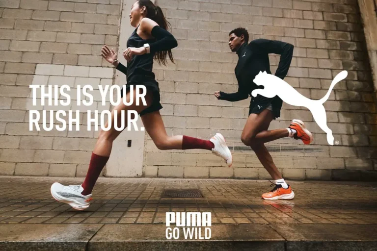 Dos corredores vestidos de ropa deportiva corren por una acera mojada junto a una pared de ladrillos beige, con el logo de PUMA y el texto “Esta es tu hora punta”, una declaración audaz en el marketing deportivo.