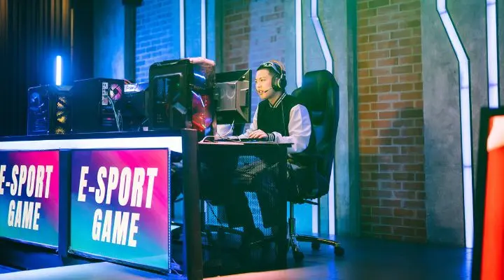 Una persona está sentada en una mesa de juego con varios monitores, con auriculares y usando un teclado y un ratón. Dos grandes pantallas muestran con claridad "Los esports", resaltando la emoción de los juegos competitivos.