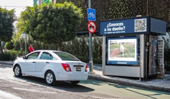 Un coche blanco se detiene cerca de una parada de autobús con la foto de un perro y un cartel con un código QR que pregunta si alguien conoce a su dueño. Cerca se ven una señal para ciclistas y árboles.