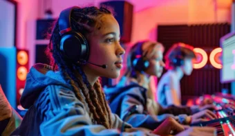 Una persona que usa auriculares y una sudadera con capucha se concentra atentamente en una pantalla de computadora en una configuración de juego, con luces de colores y equipos visibles en el fondo.