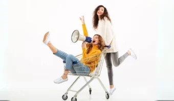 Dos mujeres se divierten con un carrito de la compra. Una está de pie detrás, sonriendo y gesticulando, mientras que la otra está sentada dentro, sosteniendo un megáfono y levantando una pierna. Ambas llevan ropa informal y zapatillas deportivas.