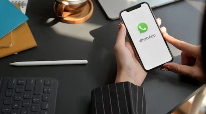 Persona sosteniendo un teléfono inteligente con la aplicación WhatsApp abierta, sentada en un escritorio con un teclado, un bloc de notas, un bolígrafo y una taza de café, mostrando la calculadora rápida de ROI