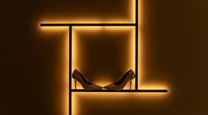Un par de zapatos de tacón alto de collabs de lujo se exhiben en un estante con líneas geométricas y una cálida iluminación sobre un fondo oscuro.