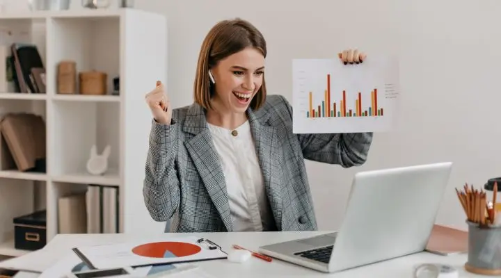 Una mujer vestida de negocios se sienta en un escritorio, sonriendo y sosteniendo un gráfico de barras, mientras mira una computadora portátil en un entorno de oficina moderno, lista para compartir verdades sobre ventas con su equipo.