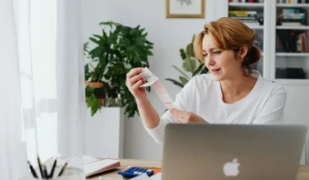 Una mujer está sentada en un escritorio con una laptop, sosteniendo y leyendo un recibo. Al fondo hay plantas, libros y material de oficina.