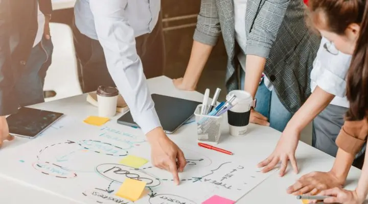 Cuatro personas de la agencia están de pie alrededor de una mesa, revisando un diagrama con notas y flechas en papel. Hay notas adhesivas, bolígrafos, una taza de café y dispositivos electrónicos dispersos por la mesa.
