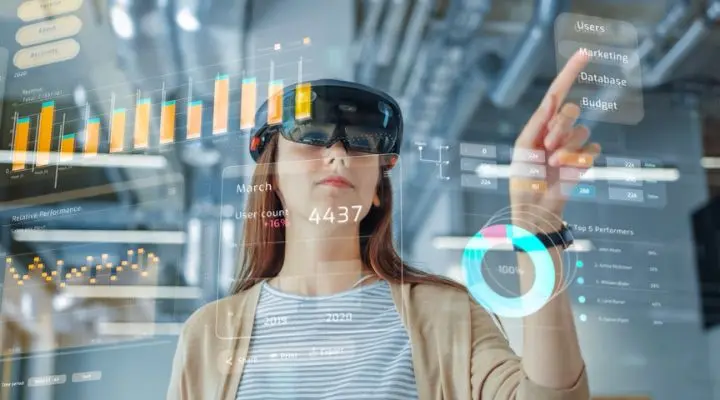 Una mujer que usa gafas de realidad aumentada interactúa con gráficos financieros virtuales y visualizaciones de datos, mostrando soluciones tecnológicas innovadoras de la agencia.