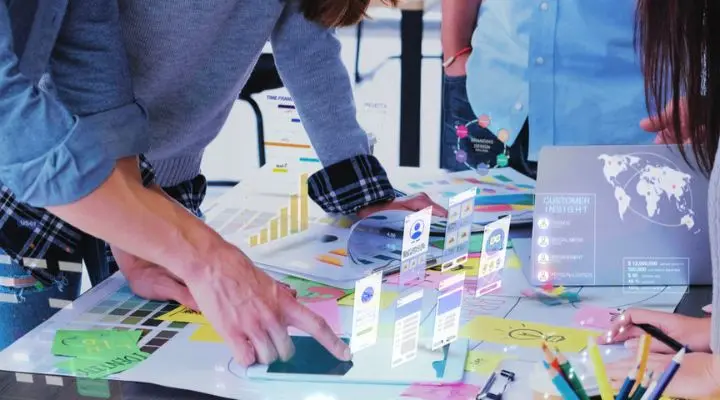 Tres personas en la agencia colaboran en un escritorio lleno de cuadros, gráficos y proyecciones de interfaces digitales, analizando datos y discutiendo el trabajo.