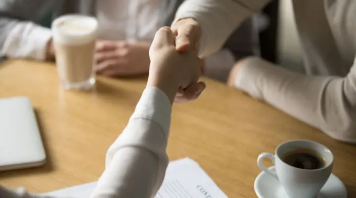 Dos personas se dan la mano sobre una mesa preparada con tazas de café, documentos y una computadora portátil cerrada, sellando un acuerdo de venta.