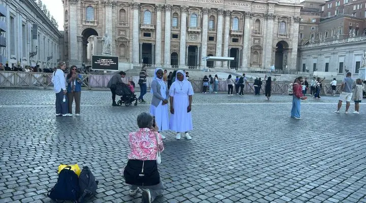 Una persona se arrodilla para tomar una fotografía de dos mujeres con hábitos blancos posando en la Plaza de San Pedro, capturando su alegre mensaje mientras otros visitantes caminan y se reúnen en el fondo.