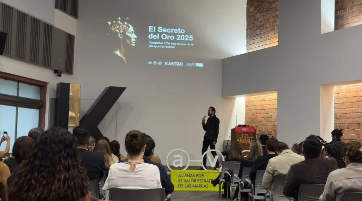 Un orador presenta "El secreto del oro 2025" a una audiencia en una moderna sala de conferencias, con una diapositiva proyectada y una gran letra "K" como parte de la decoración.