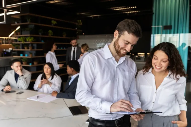 Dos colegas con camisas blancas revisan un portapapeles en una mesa de oficina moderna, encarnando el esfuerzo por humanizar la marca, mientras varios otros se reúnen y conversan en el fondo.