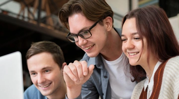 Tres adultos jóvenes se sientan juntos, sonriendo y mirando una pantalla de computadora en un ambiente interior informal, tal vez investigando las agencias para su próximo gran proyecto.