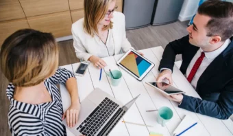 Tres personas se sientan en una mesa con computadoras portátiles, tabletas, teléfonos y tazas de café, participando en una discusión en un entorno de oficina moderno.
