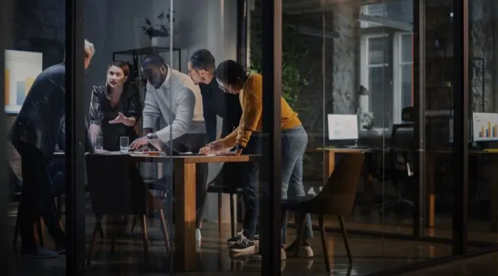 Cinco personas de la agencia están de pie alrededor de una mesa en una oficina moderna, revisando documentos y una laptop, enfrascados en una conversación. Al fondo se ven monitores con gráficos.
