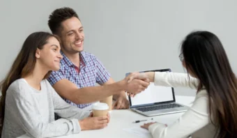 Un hombre y una mujer se sientan en un escritorio con café, estrechando la mano de una empresaria frente a ellos; hay una computadora portátil y papeles sobre la mesa, discutiendo detalles sobre programas de lealtad.