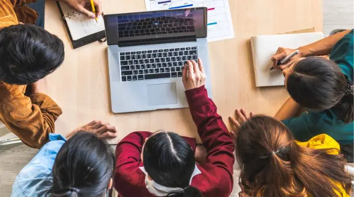 Cinco personas se sientan alrededor de una mesa en la agencia, mirando una computadora portátil y tomando notas en papel, participando en una sesión de estudio o trabajo colaborativo.