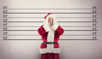Una persona vestida de Papá Noel se encuentra frente a un muro de medición de altura, con aspecto sorprendido, como si estuviera en una rueda de reconocimiento policial.