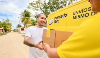 Una persona recibe un paquete de un repartidor de Mercado Libre frente a una camioneta amarilla en un día soleado.