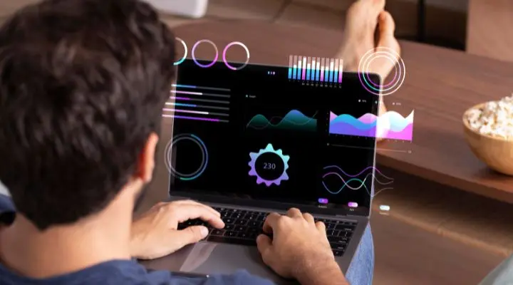 Una persona trabajando en una computadora portátil que muestra gráficos y cuadros coloridos para la medición del marketing, con los pies apoyados sobre una mesa y un tazón de palomitas de maíz cerca.
