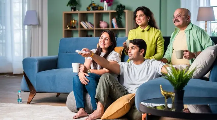 Cuatro personas están sentadas en una sala de estar, formando audiencias relajadas alrededor del televisor (dos en un sofá azul, una en el suelo), y todas parecen comprometidas y contentas juntas.